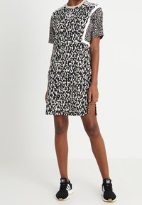 Žena v šatech s krátkým rukávem, černých a béžových s vzorem a bílým pruhovaným detailem, doplněná černo-bílými teniskami Adidas.