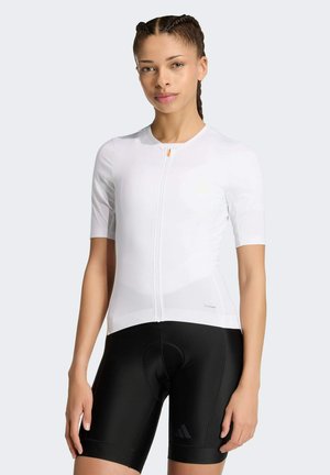 Młoda kobieta ubrana w białą koszulkę kolarską z krótkim rękawem i zamkiem oraz czarne wyściełane spodenki kolarskie, stojąca na jednolitym tle.