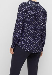 Langärmlige Bluse mit einem dunkelblauen Hintergrund, die ein lila und elfenbeinfarbenes abstraktes Muster zeigt, aus weichem Stoff besteht und einen geknöpften Halsausschnitt hat.