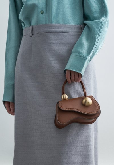 Nainen, jolla on päällään vaalean teal-pusero ja harmaat hameet, pitää kädessään pientä ruskeaa käsilaukkua, jossa on kultaisia pyöreitä koristeita.