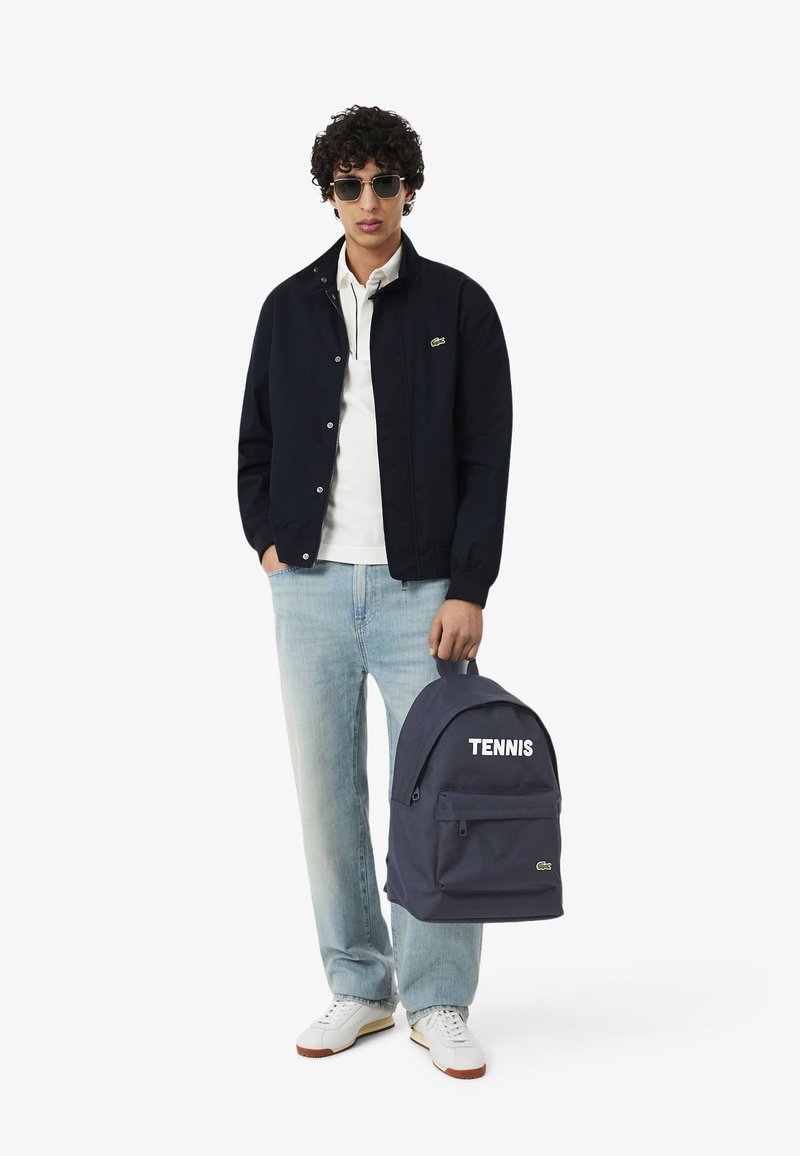 Giovane uomo con occhiali da sole, giacca blu scuro, polo bianca, jeans chiari, sneaker bianche, che tiene uno zaino blu scuro con l'etichetta "TENNIS" su sfondo bianco.