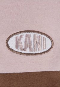 Brodert logo "KANI" i beige, på et lys rosa stoff med et brunt stripet mønster. Teksturen er glatt og strikket.