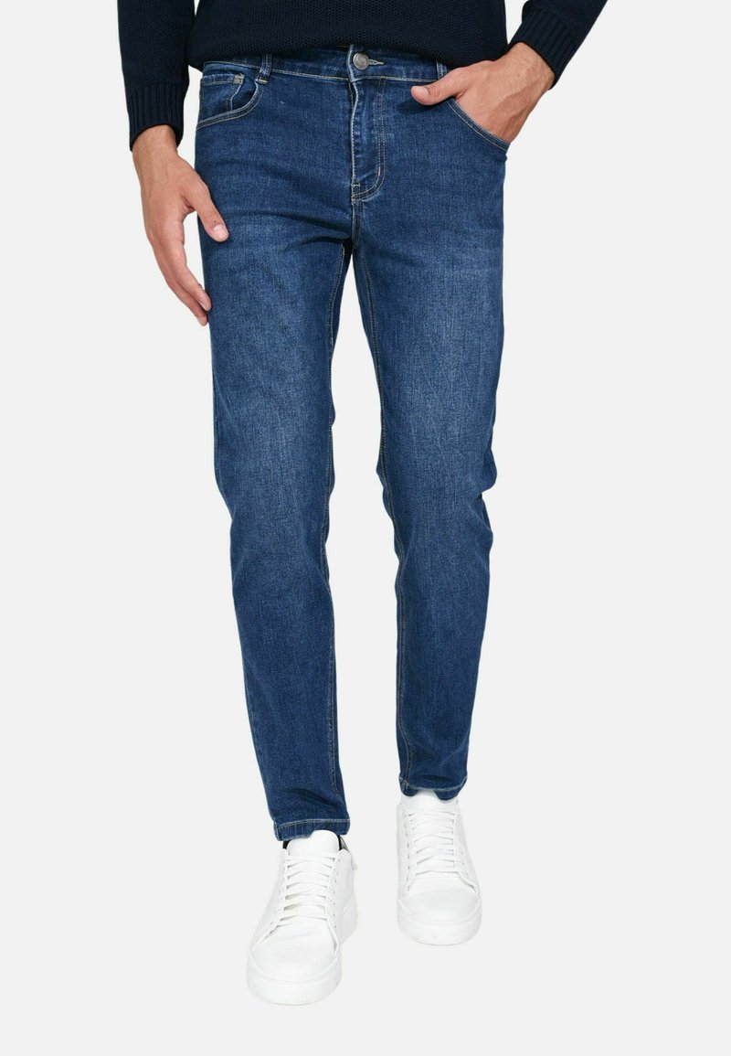 Jeans in denim blu scuro con vestibilità slim, design a cinque tasche e zip. Indossati con sneakers bianche per un look casual.