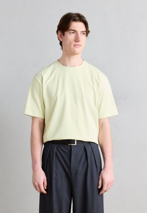 Jeune homme aux cheveux bruns portant un t-shirt jaune pâle rentré dans un pantalon plissé gris foncé taille haute, sur un fond uni.
