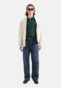 Kremowa kurtka na zamek, noszona na zielonym podkoszulku, połączona z niebieskimi dżinsami i czarnymi butami, uzupełniona ciemnymi okularami przeciwsłonecznymi.