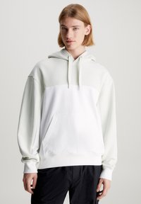 Sudadera con capucha verde claro y blanca con un panel frontal blanco sólido, bolsillo canguro y puños acanalados. Textura de tela suave en todo su diseño.