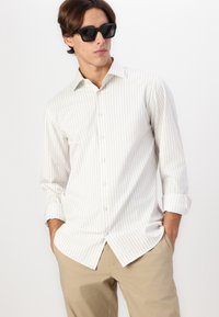 Camisa de manga longa, com botões, em riscas verticais claras de bege e branco; tecido de algodão, colarinho clássico, punhos enrolados, usada com óculos de sol pretos.