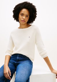 Κρεμ πλεκτό πουλόβερ με λαιμόκοψη βαρκούλα, μακριά ραγκλάν μανίκια και μικρό κεντητό λογότυπο. Συνδυασμένο με μπλε τζιν παντελόνι. Λείος υφής.