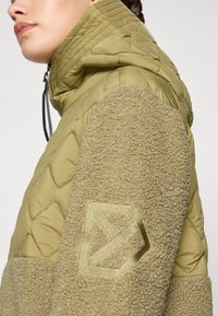 Olivengrønn zip-jakke med quiltet stoff og beige, teksturert fleece-ermene. Har et dekorativt logo-merke på ermet.