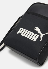 Czarna torba na ramię z dużym białym logo "PUMA" oraz charakterystycznym sylwetką kota. Teksturowany materiał o płaskim kształcie i regulowanym pasku.