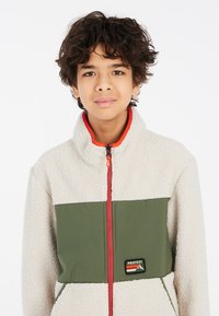 Chaqueta de forro polar con un cuerpo color crema y un panel verde, que presenta una cremallera roja, un cuello alto y una placa de logo "PROTEST" en el frontal.