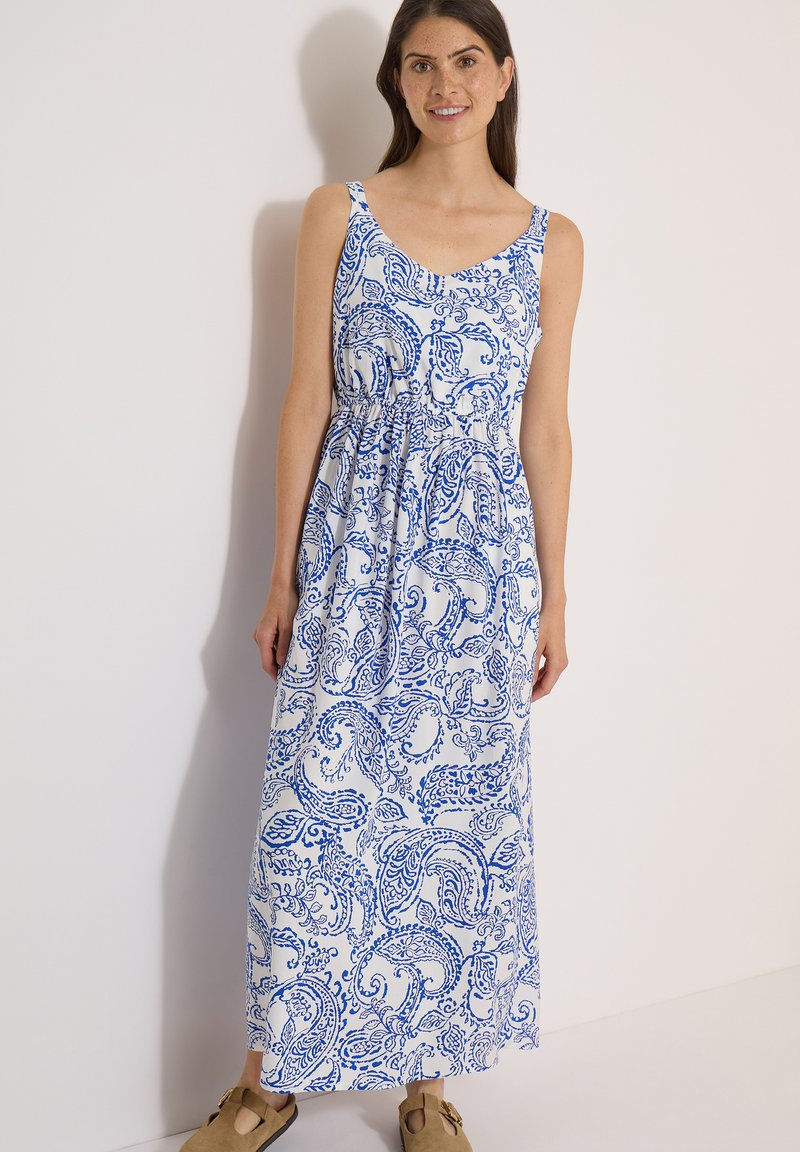 Femme debout, souriante, vêtue d'une robe longue blanche sans manches à imprimé cachemire bleu et de sandales beiges, sur un fond clair et uni.