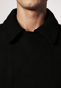 Abrigo de lana negro con cuello abierto, que presenta una textura suave y líneas limpias, costuras visibles a lo largo de la tapeta y un diseño minimalista.