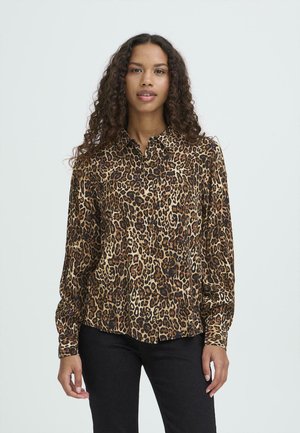 Donna con capelli lunghi e ricci, che indossa una camicetta marrone e nera con stampa leopardata e pantaloni neri, in piedi davanti a uno sfondo semplice.