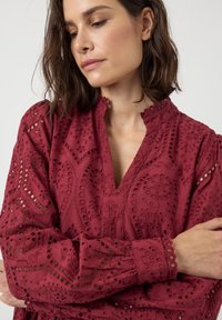 Burgunderfarget brodert bluse med intrikate utskjæringsmønstre, V-hals og bølgede mansjetter. Laget av lettvektig stoff med en teksturert overflate.