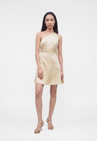 Femme aux longs cheveux foncés portant une mini-robe en satin crème à une épaule et des sandales à talons hauts dorées, debout sur un fond blanc uni.