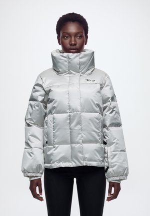 Modèle portant une doudoune matelassée argentée et brillante, avec un col haut rembourré, des poches avant et un pantalon noir, sur un fond blanc uni.
