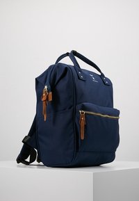 Námořnická modrá plátěná batoh s hnědými koženými akcenty, s přední kapsou na zip a dvojitými ramenními popruhy. Má strukturovaný tvar.