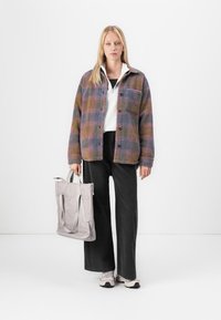 Giacca over size a quadri nei toni viola, marrone e grigio. Indossata sopra una maglietta bianca e pantaloni neri. Borsa tote grigia con manici e sneakers chiare.