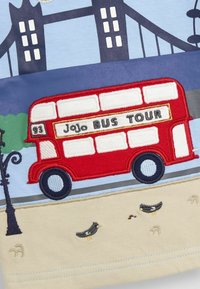 Piros hímzett busz, rajta a "JoJo BUS TOUR" felirattal egy kék háttéren, mellette egy bézs színű terület, amelyen apró madarak és egy lámpaoszlop motívuma látható.