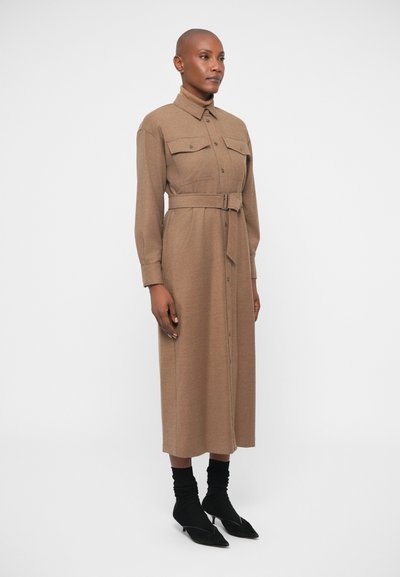 Vestido camisola de manga longa castanho em tecido texturizado, com uma cintura cingida por cinto, frente com botões e dois bolsos peitorais. Usado com meias e sapatos pretos.