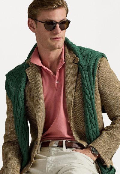 Blazer maro tip herringbone, cămașă polo roz, pulover verde tricotat aruncat pe umăr; accesorizat cu ochelari de soare și un ceas.