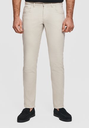 Beżowe spodnie o dopasowanym kroju noszone przez osobę w czarnych mokasynach, widok z przodu od talii do kostek na jednolitym tle.