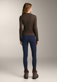 Smeđa dugačka majica s dugim rukavima, prilagođenog kroja i rebrastom teksturom, uparena s tamnoplava tajicama i smeškim čizmama do gležnja s krznom.