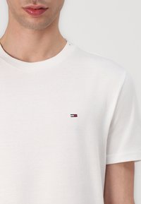 Tricou alb din bumbac cu guler rotund, având un mic logo brodat în roșu, alb și albastru pe partea stângă a pieptului. Textură moale, mâneci scurte.