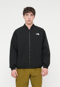 Μαύρο μπουφάν με φερμουάρ, γιακά σε όρθια στάση, ελαστικά μανίκια, και μικρό λευκό λογότυπο στο αριστερό στήθος. Ελαφρύ, με λεία υφή, βασικό σχέδιο.