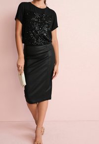 Haut noir à manches courtes avec un devant en sequins, associé à une jupe crayon en cuir noir ajustée. Une pochette beige complète la tenue.