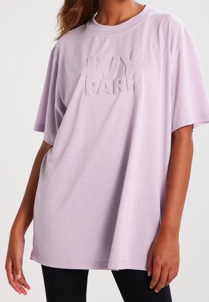 Kobieta w luźnym, jasnofioletowym t-shircie z wypukłym tekstem "IVY PARK" na piersi i czarnych legginsach, stojąca na jednolitym tle.