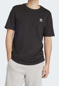 Svart bomullströja med korta ärmar och rund halsringning, med en vit Adidas-logotyp på vänster bröst. Kombination med gråa sweatpants.