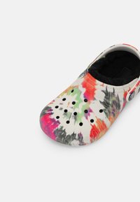 Chaussure style sabot avec motif tie-dye en rouge, vert, violet et noir, présentant des trous de ventilation et une doublure en polaire noire à l'intérieur.