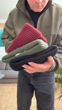 Négy kötött sapka egymásra rakva: piros, oliva zöld és fekete. Bordás textúra, puha anyag. Kézben tartva, világos háttérrel és bútorokkal.