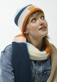 Gehaakte muts met oranje, blauwe en crèmekleurige strepen, voorzien van een vierkante tan patch. Bijpassende dikke sjaal in vergelijkbare kleuren en texturen.