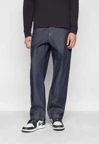 Jeans de mezclilla azul oscuro con un corte relajado, con costuras en contraste y un diseño clásico de cinco bolsillos, combinados con zapatillas negras y blancas.