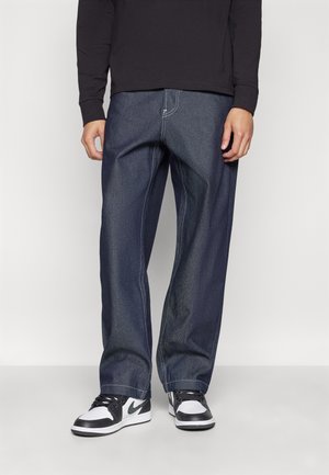 Jeans de mezclilla azul oscuro con un corte relajado, con costuras en contraste y un diseño clásico de cinco bolsillos, combinados con zapatillas negras y blancas.