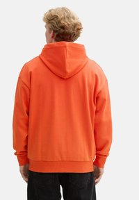 Sudadera naranja brillante con una gran capucha, puños y dobladillo acanalados, hecha de material suave, que presenta un corte relajado y un diseño minimalista.
