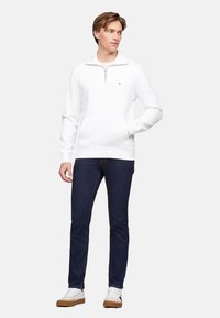 Sudadera blanca de cuello con media cremallera y bolsillo frontal; combinada con jeans azul oscuro y zapatillas blancas; estilo casual.