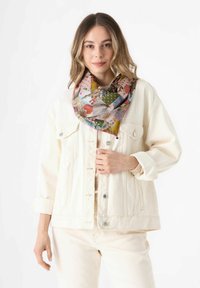 Multicolore zijden sjaal met een patchworkontwerp met bloem- en geometrische patronen, om de nek gedrapeerd over een witte denimjack.