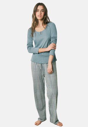 Nainen seisoo paljain jaloin, päällään vaaleansininen pitkähihainen paita ja väljät raidalliset pyjamahousut, kädet ristissä ja rentona.