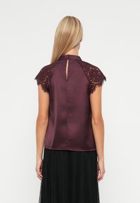 Top in satin bordeaux con maniche corte in pizzo, schiena a chiave di violino e orlo arrotondato. Tessuto morbido con dettagli in pizzo intricato sulle spalle.