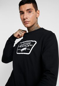 Sudadera negra de manga larga con un logo gráfico blanco y el texto "VANS NY Desde 1966" en el frente. Textura suave.