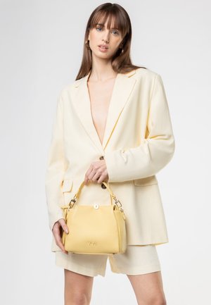 Donna che indossa un blazer crema oversize e pantaloncini, tiene una piccola borsa gialla con accessori dorati e logo "V73".