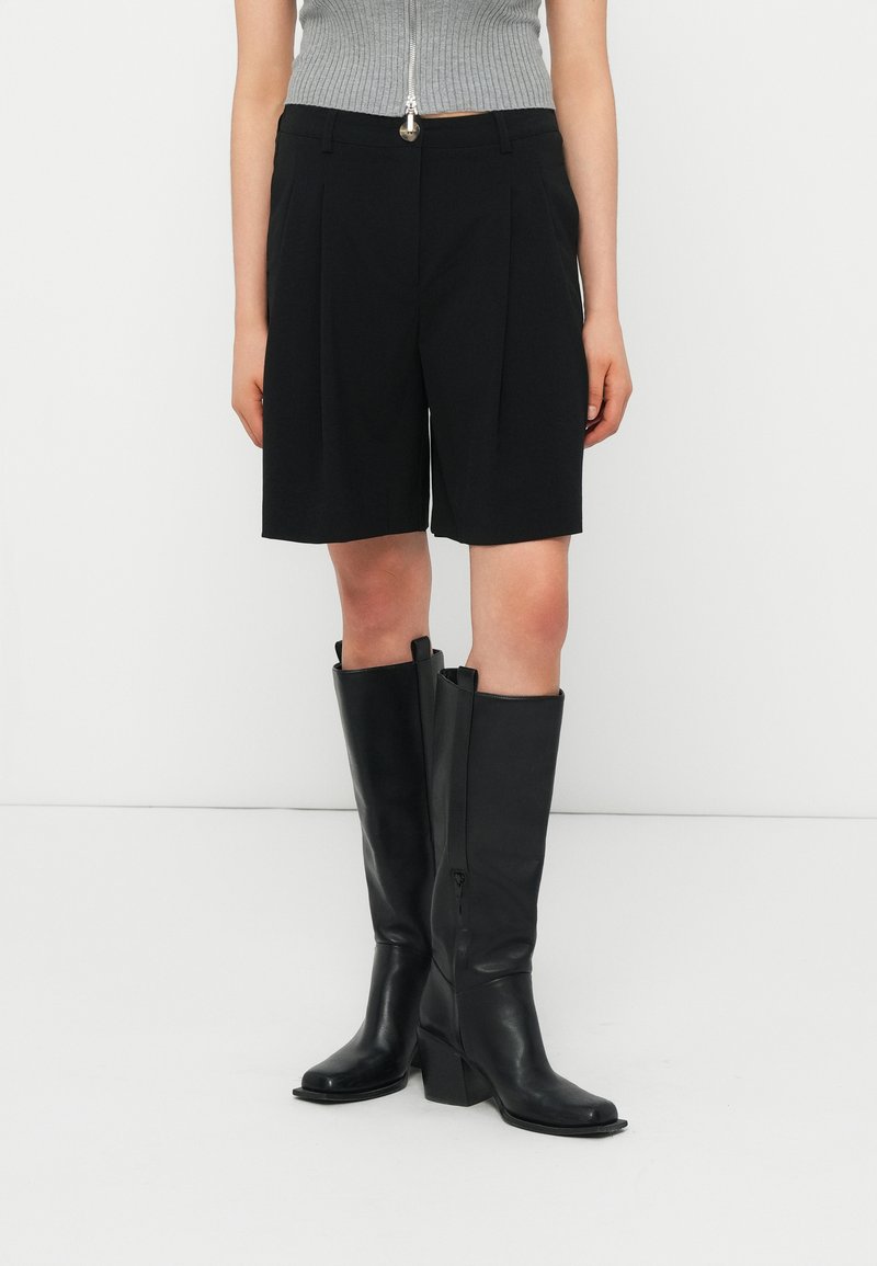 Personne portant des bottes noires montantes jusqu'aux genoux, un short noir arrivant aux genoux, et un haut gris sans manches à côtes avec une fermeture éclair à l'avant.