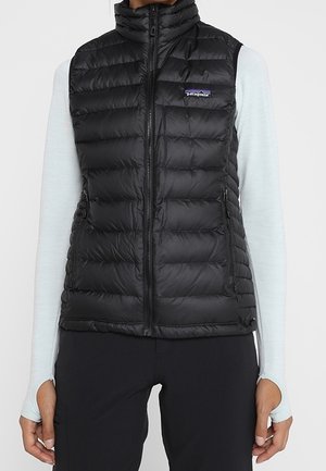Femme portant une veste matelassée noire Patagonia par-dessus un haut gris clair à manches longues avec des trous pour les pouces et un pantalon noir.
