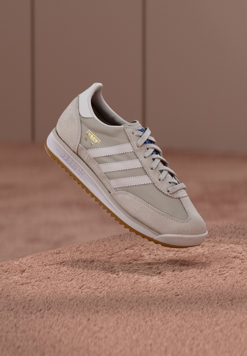 Beige joggesko med overdel i semsket skinn og mesh, hvite tre striper, såle i gummigummi, blå lisser og gullfarget logo. Klassisk retrodesign.