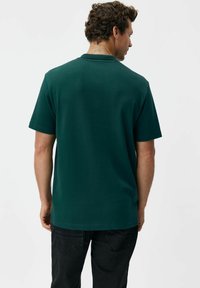 Ciemnozielony t-shirt z krótkim rękawem wykonany z bawełny, z okrągłym dekoltem i luźnym krojem. Tył jest gładki, bez widocznych logo czy wzorów.