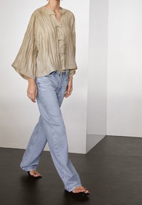 Blouse ample plissée beige clair avec col en V et fermetures à boutons, associée à un jean large bleu clair et des sandales noires ouvertes à l'avant.